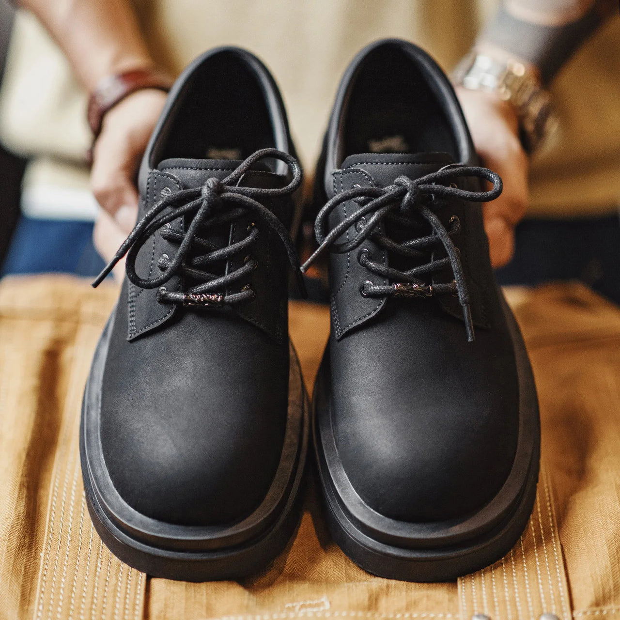 A pair of black leather lace-up shoes with chunky soles, held by hands over a tan fabric surface.