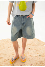A person standing on sand wearing light-wash denim shorts, a gray graphic T-shirt, and yellow flip-flops.