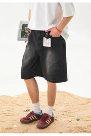A person wearing black denim shorts, a white t-shirt, white socks, and burgundy sneakers with gold stripes, standing on a sandy surface against a white background.