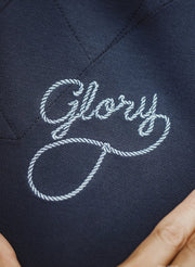 Close-up detail shot of a dark navy garment with white embroidered script reading "Glory" on the chest, with fingers holding the fabric at the bottom and the word "DETAILS" at the top.