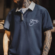 A close-up of a man wearing a navy blue short-sleeve polo shirt with a white collar, featuring a white embroidered script logo on the chest that reads 'Glory'. The man has a full beard, tattoos on his neck and arms, and is wearing a silver pendant necklace.