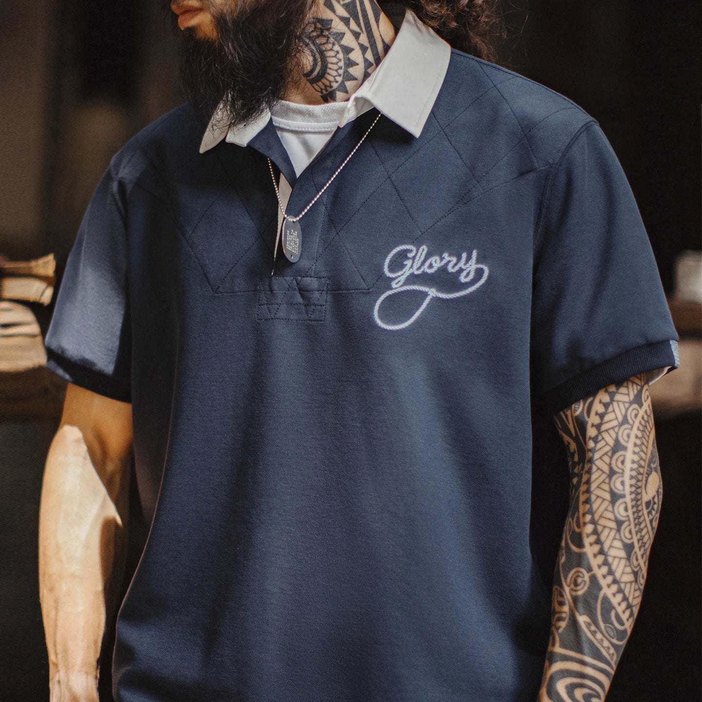 A close-up of a man wearing a navy blue short-sleeve polo shirt with a white collar, featuring a white embroidered script logo on the chest that reads 'Glory'. The man has a full beard, tattoos on his neck and arms, and is wearing a silver pendant necklace.