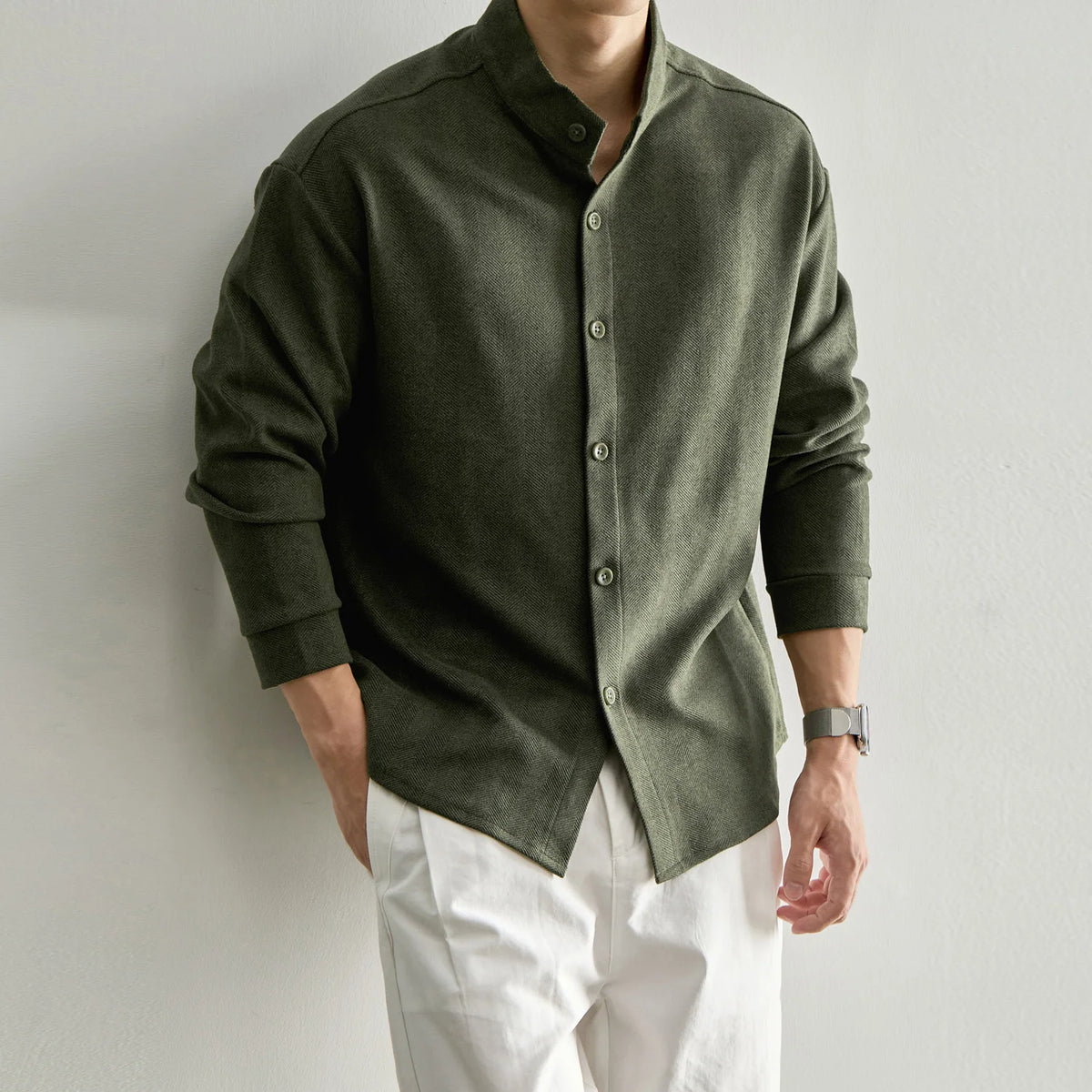 A man wearing a dark olive green long-sleeve button-up shirt with a mandarin collar and white pants, standing against a plain light gray wall.