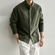 A man wearing a dark olive green long-sleeve button-up shirt with a mandarin collar and white pants, standing against a plain light gray wall.