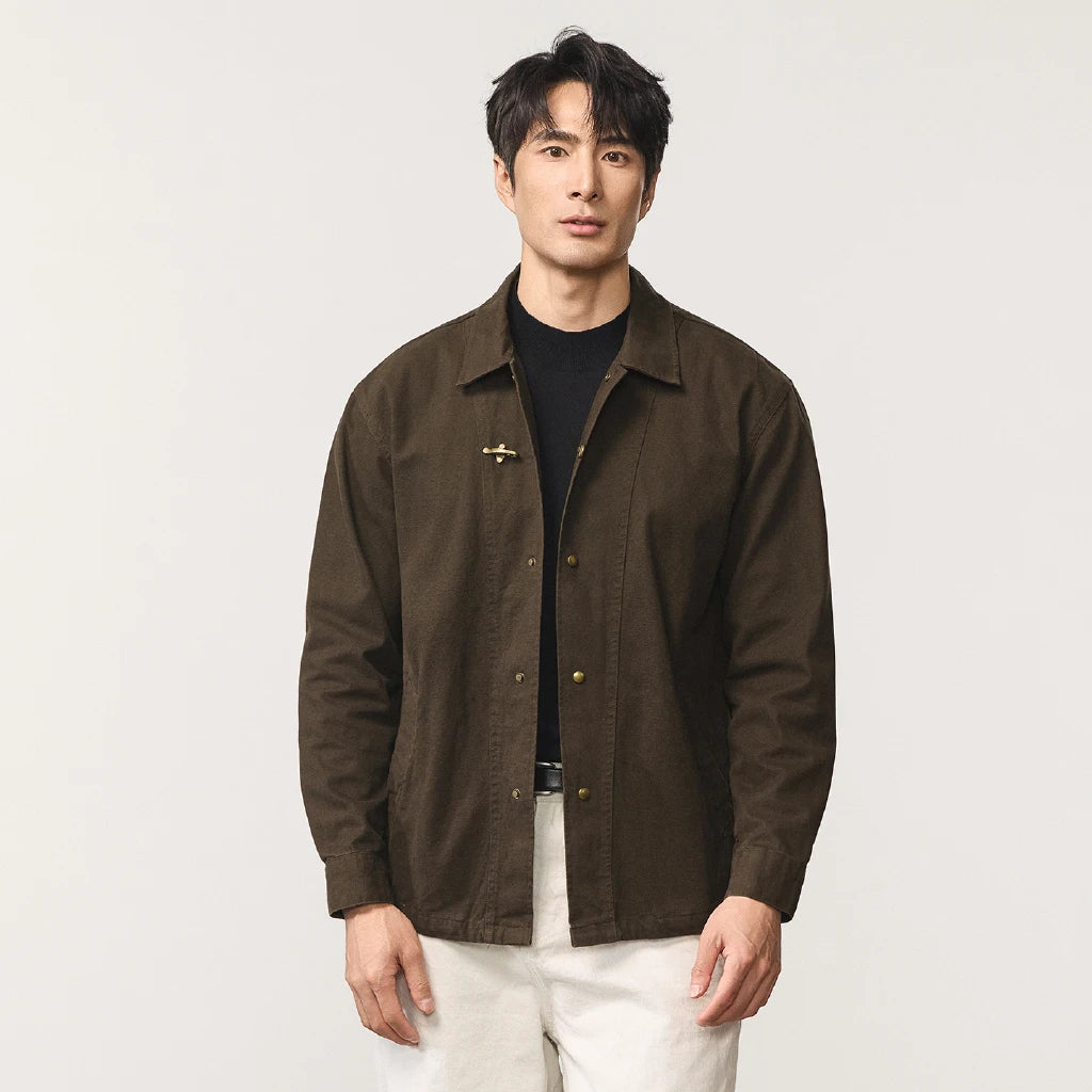 A man wearing a dark brown button-up overshirt with a small cross-shaped metal accent on the chest, layered over a black t-shirt and paired with off-white trousers, photographed against a light gray studio background.