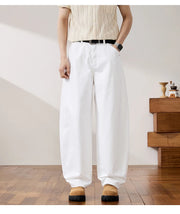 A person wearing loose white wide-leg pants with a black belt, paired with a cream striped short-sleeve top and tan shoes, standing in a minimalist room with a wooden floor and side table.