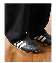 Black slip-on shoes with three white stripes, shown being worn with black pants on a wooden floor. 