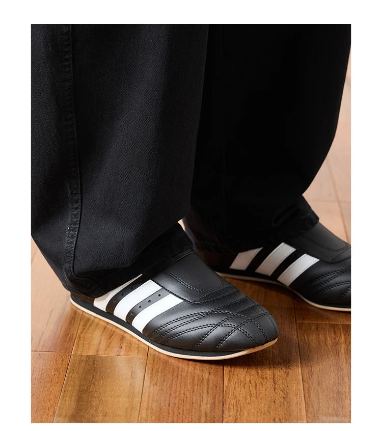 Black slip-on shoes with three white stripes, shown being worn with black pants on a wooden floor. 