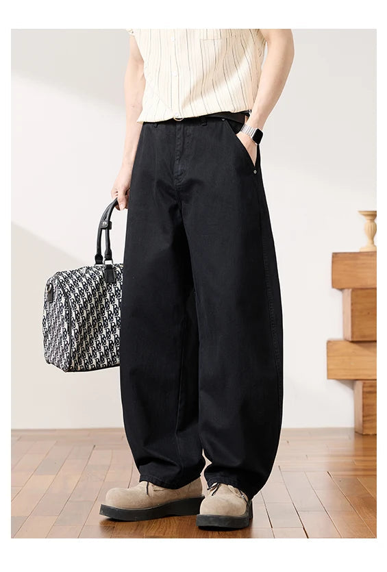 A model wearing black wide-leg baggy pants, a cream ribbed tank top, and beige shoes, holding a black and white patterned handbag in a minimalist indoor setting.