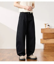 A person wearing loose black wide-leg pants with a white belt, paired with a cream ribbed top and beige shoes, standing in a bright indoor room with wood flooring and a wooden side table.