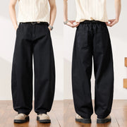 Two views of a person modeling loose black wide-leg pants with a relaxed straight-to-sweatdrop silhouette, paired with a cream striped shirt and beige shoes, shown against a white wall and wood floor.