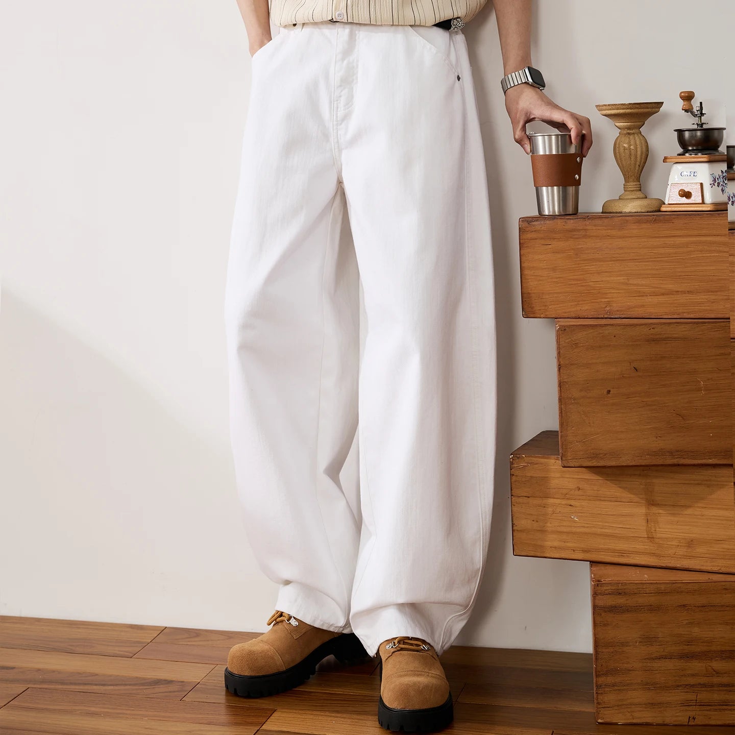 White wide-leg pants worn by a model standing in a room with wooden stairs and a coffee grinder, holding a metal cup.