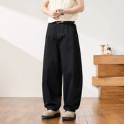 A person wearing loose black wide-leg pants with a black belt, paired with a cream ribbed short-sleeve top and beige shoes, standing in a bright indoor room with wood flooring and a wooden side table.