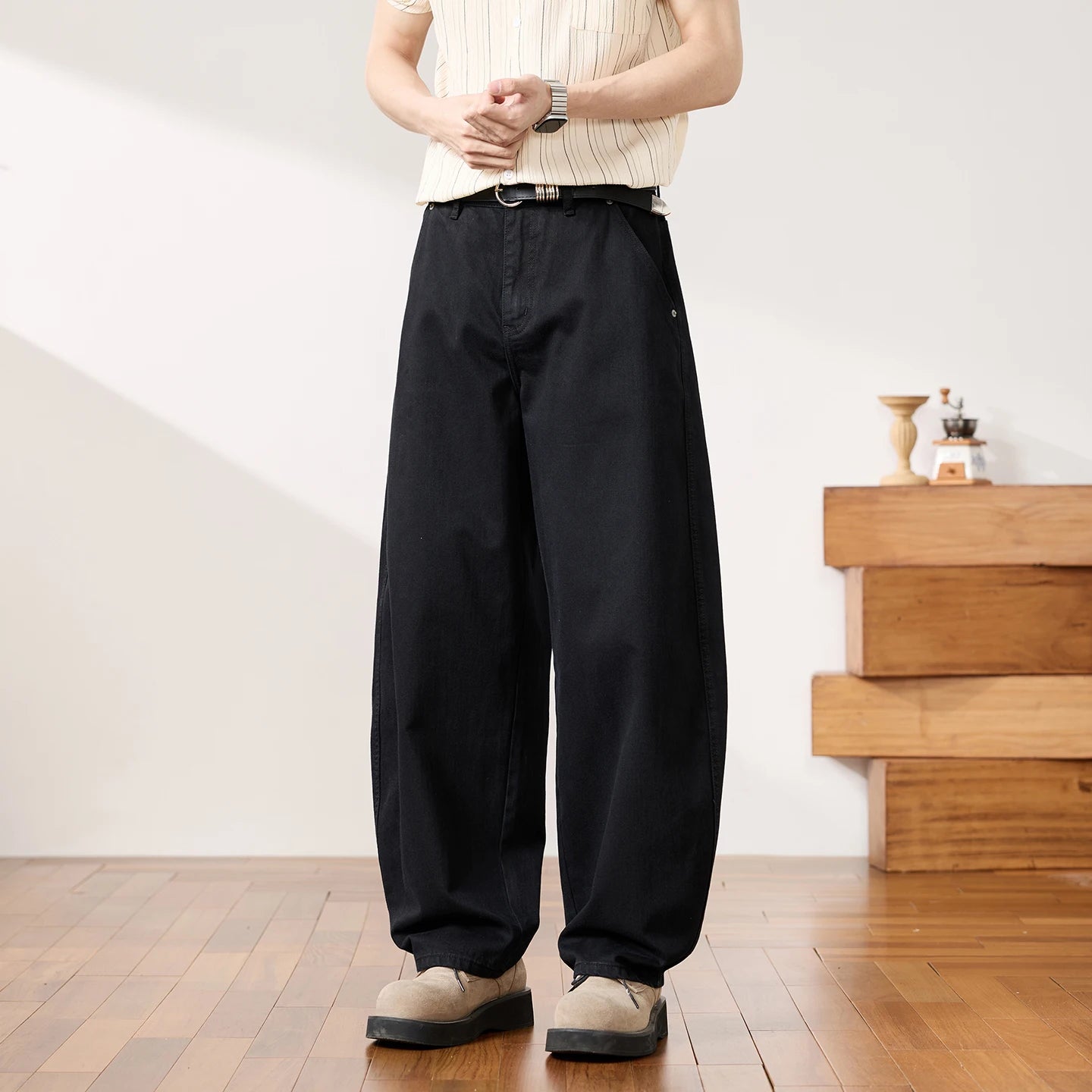 A person wearing loose black wide-leg pants with a black belt, paired with a cream ribbed short-sleeve top and beige shoes, standing in a bright indoor room with wood flooring and a wooden side table.
