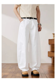 A person wearing loose white wide-leg pants with a black belt, paired with a cream short-sleeve top and tan shoes, standing in an indoor room with wood flooring and a wooden side table.