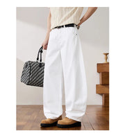 A person wearing white wide-leg trousers, a cream ribbed top, and tan suede loafers, holding a black-and-white patterned handbag in a styled indoor room with wood flooring.