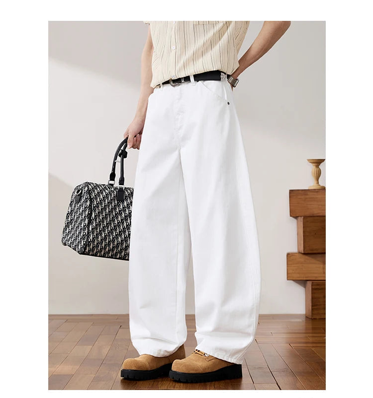 A person wearing white wide-leg trousers, a cream ribbed top, and tan suede loafers, holding a black-and-white patterned handbag in a styled indoor room with wood flooring.