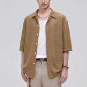A model wearing a textured, short-sleeve button-up knit shirt in a warm camel brown over a white t-shirt and beige trousers, shot against a solid light gray background.