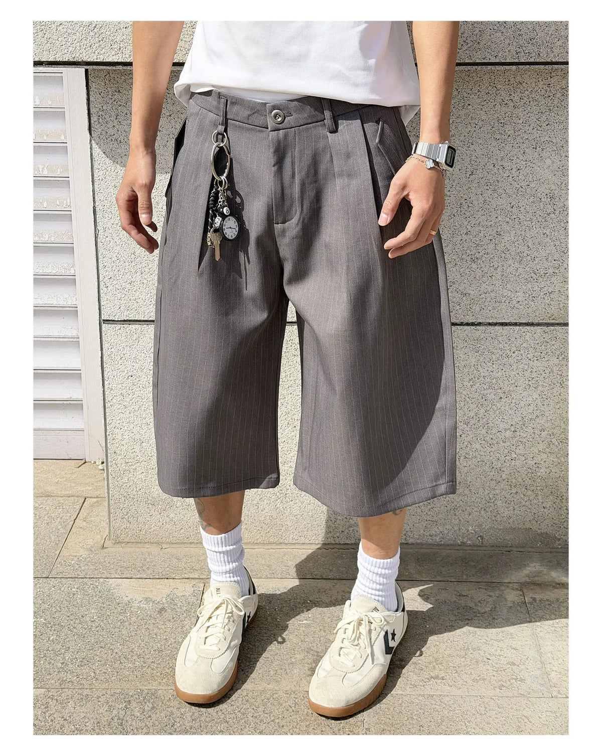 A person wearing loose, wide-leg gray shorts with a subtle vertical stripe pattern, styled with a white t-shirt, white socks, and off-white sneakers against a gray stone wall.