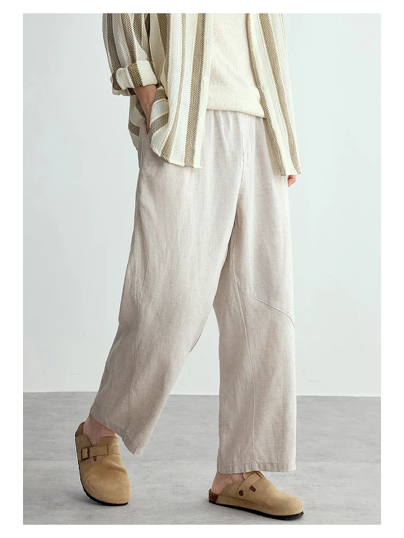 A person wearing loose, wide-leg linen pants in a light beige color, paired with a cream and beige striped shirt and tan slip-on clogs, shown from the waist down against a neutral studio background.