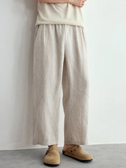 A person wearing loose, wide-leg linen trousers in a light beige color, paired with a matching cream top and tan suede clogs, standing against a plain white wall.