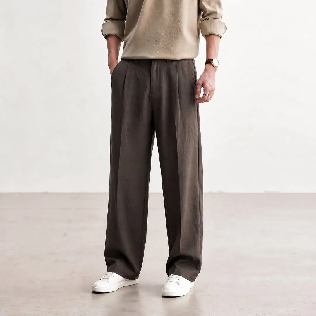 A person wearing dark brown wide-leg linen trousers, a beige knit sweater, and white sneakers, standing in a minimalist studio setting.