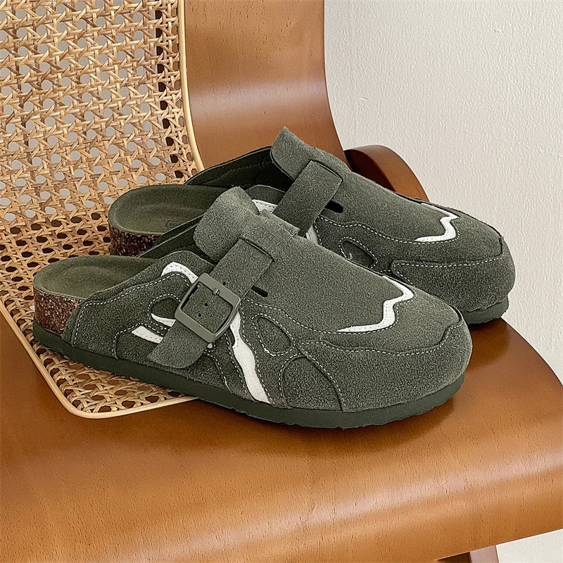 A pair of dark olive green clog-style slip-on shoes with white wavy stripe accents and dark buckles, displayed on a brown leather chair next to a woven cane seat.