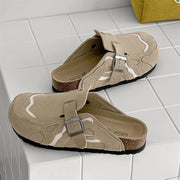 A pair of beige clog-style slide sandals with adjustable straps, silver buckles, cork midsoles, and black outsoles, displayed on a white tiled surface.