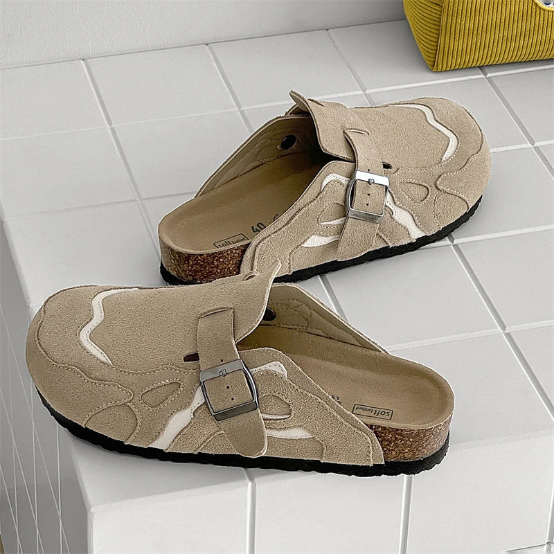 A pair of beige clog-style slide sandals with adjustable straps, silver buckles, cork midsoles, and black outsoles, displayed on a white tiled surface.
