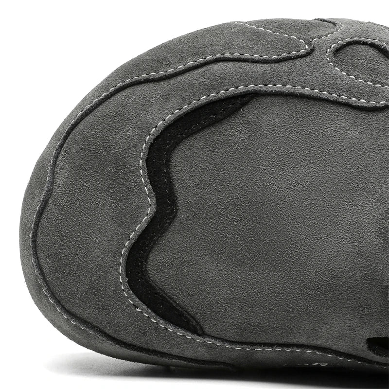 Close-up of a dark gray suede shoe upper with decorative stitching and a wavy black accent panel, photographed on a white background.