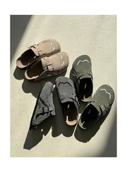 Four slip-on shoes in neutral tones arranged on a textured light surface with strong sunlight and shadows. The shoes include taupe, dark gray, and olive green versions, all with matching strap closures and decorative white line accents.