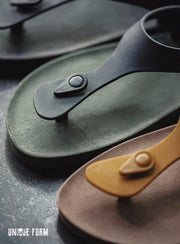 Three minimalist thong sandals with circular snap closures are arranged diagonally on a dark textured surface. The sandals are in black, muted olive green, and tan.