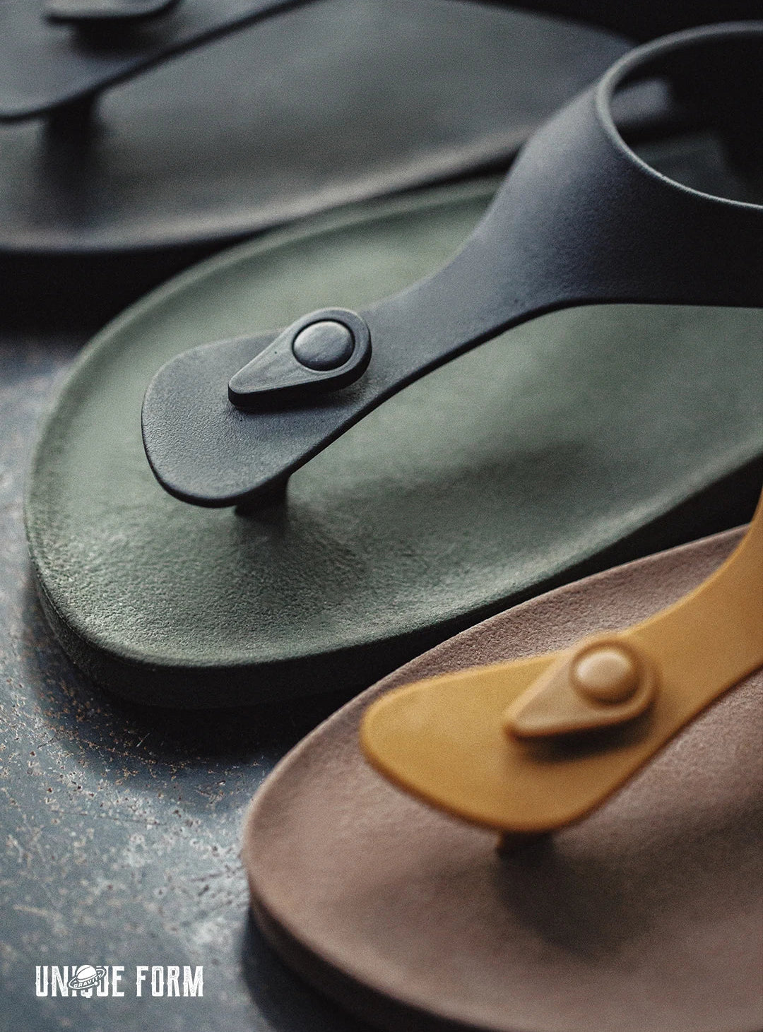 Three minimalist thong sandals with circular snap closures are arranged diagonally on a dark textured surface. The sandals are in black, muted olive green, and tan.