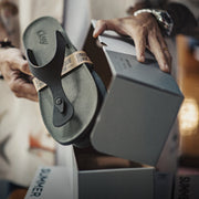 A person holds a dark olive green flip-flop sandal with a tan strap printed with 'UNIQUE FORMULA' next to an open gray shoebox. 