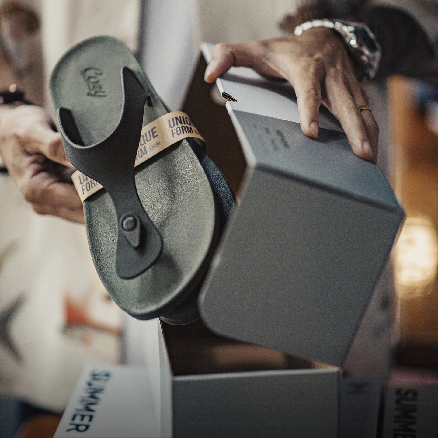 A person holds a dark olive green flip-flop sandal with a tan strap printed with 'UNIQUE FORMULA' next to an open gray shoebox. 