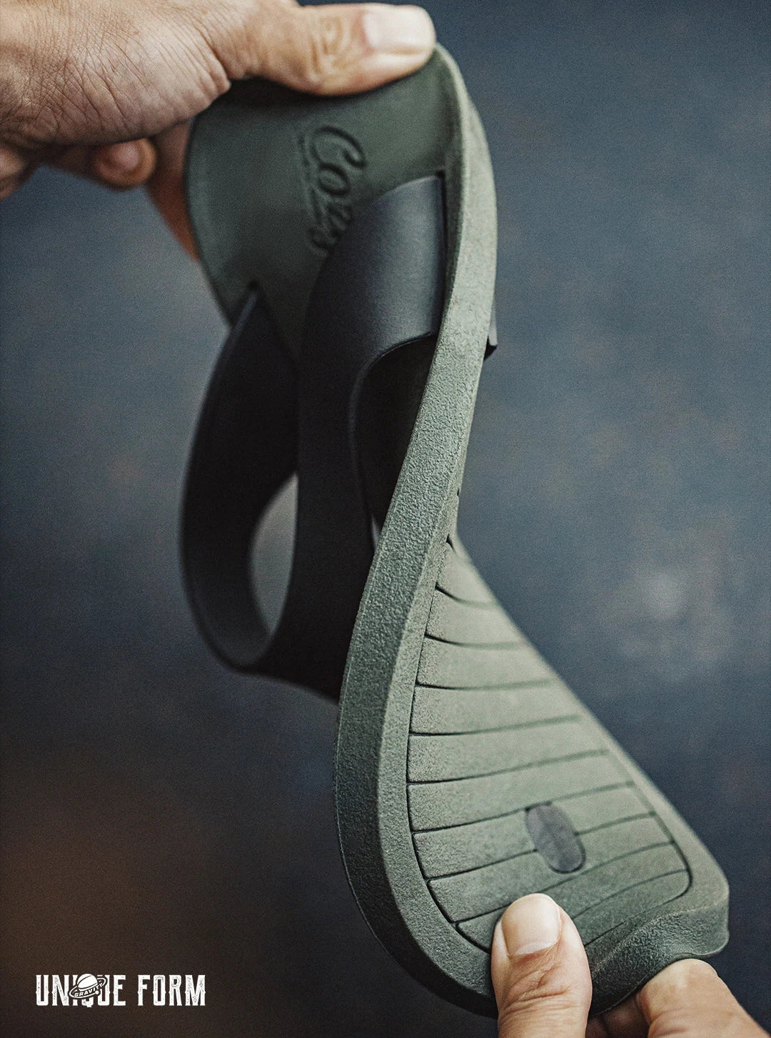 A close-up of a person holding a flexible Cozi flip-flop sandal to show its shape and sole, with the brand name 'Cozy' embossed on the footbed. The background is a dark, textured studio backdrop.