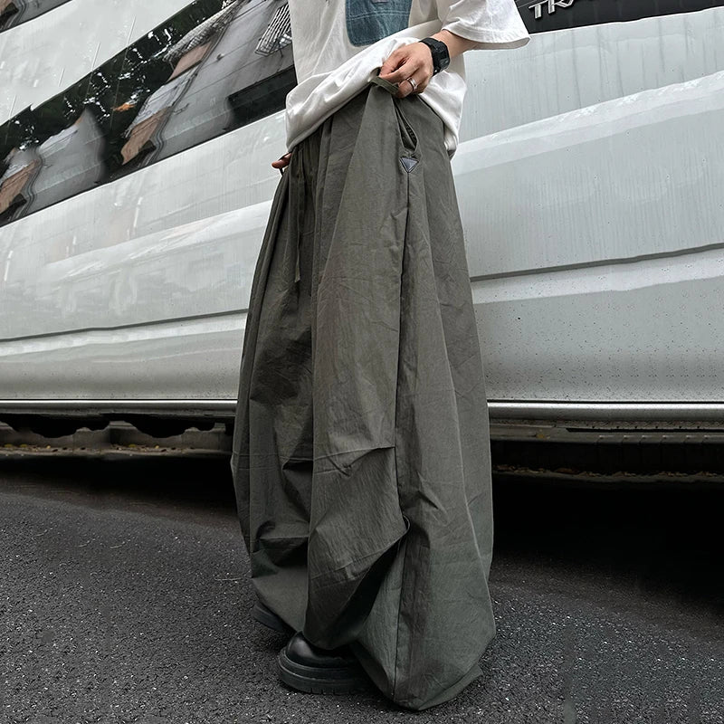 A person wearing very wide-leg, loose-fit dark gray cargo pants and a white shirt, standing on asphalt next to a white vehicle.