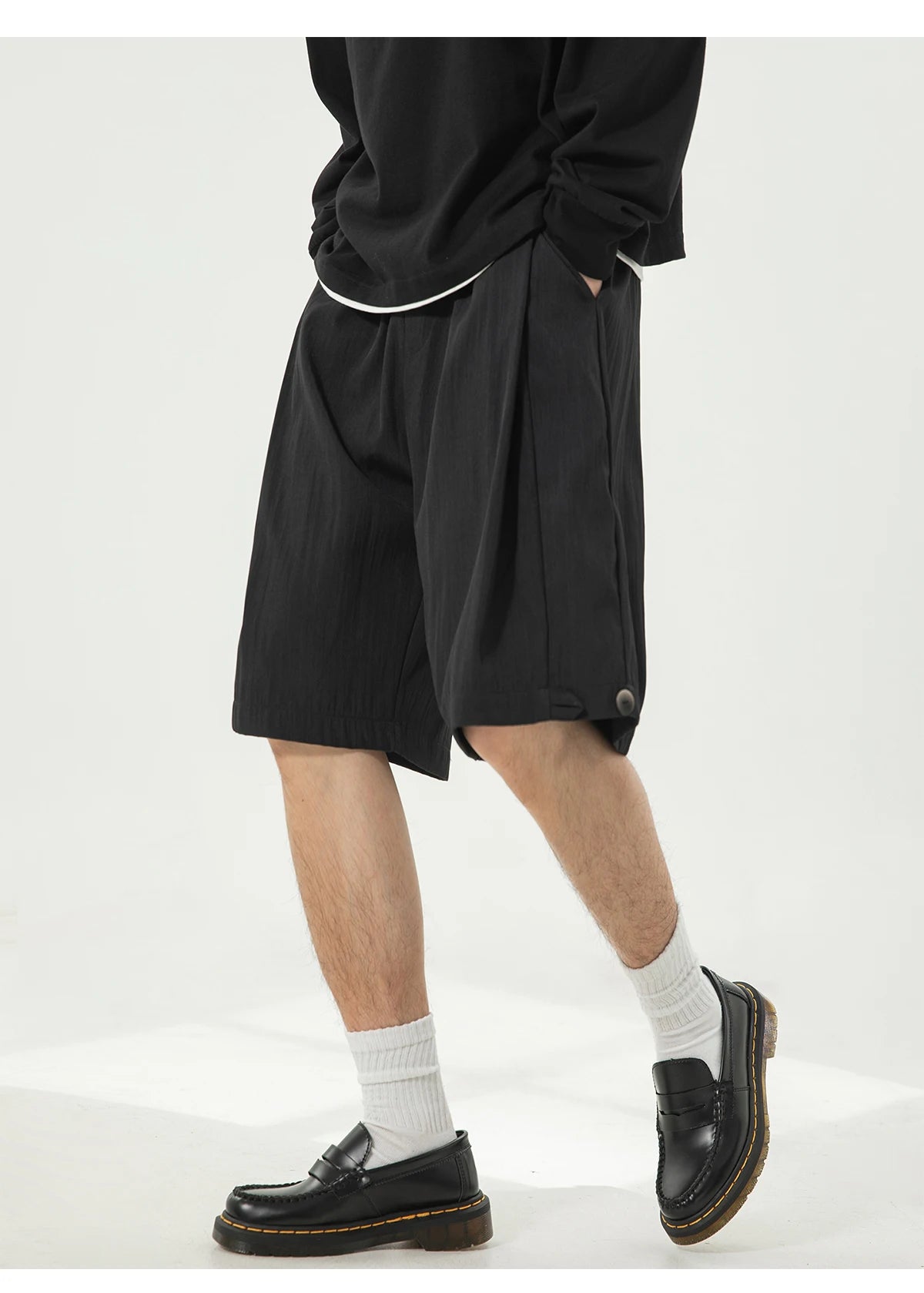 A person wearing black pleated shorts, white socks, and black leather loafers, shown from the waist down against a plain light background.
