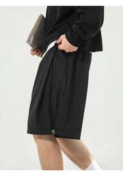 A person wearing loose black shorts and a black long-sleeve top, holding a gray clutch, photographed against a plain light background.