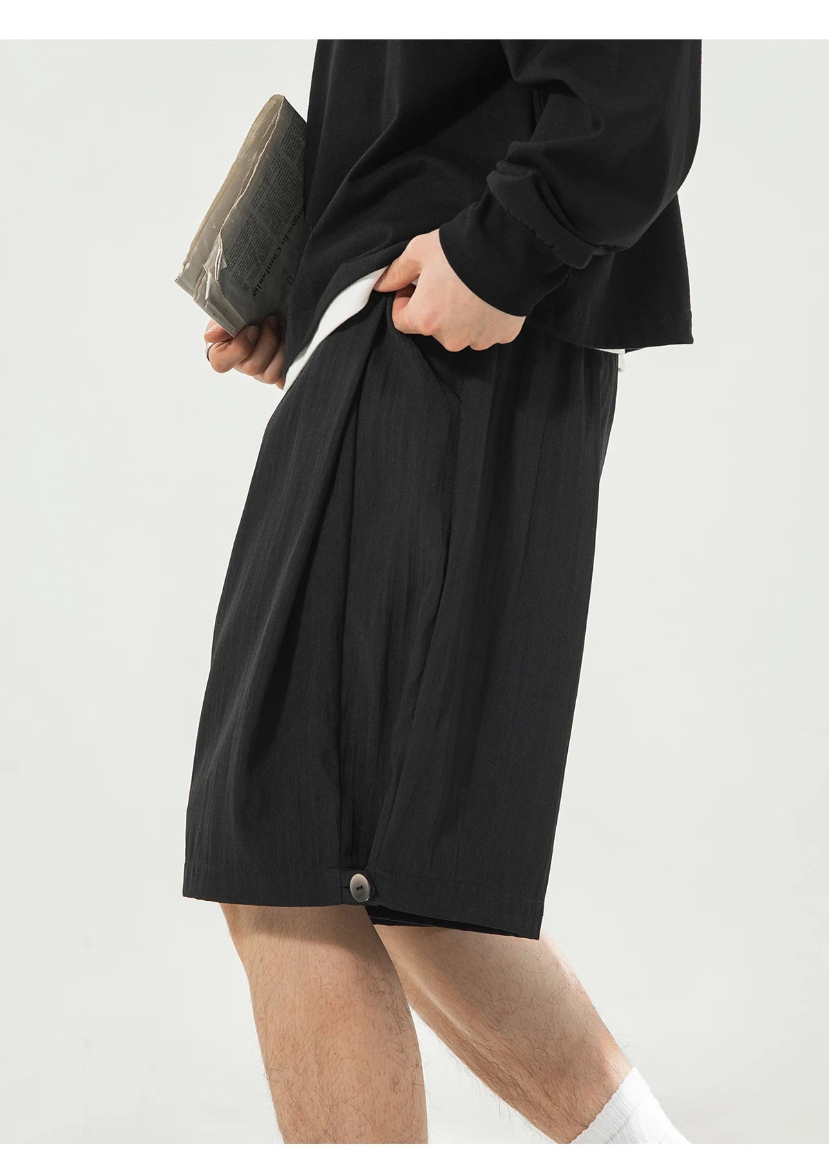 A person wearing loose black shorts and a black long-sleeve top, holding a gray clutch, photographed against a plain light background.