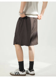 A person wearing dark brown pleated shorts, a light gray t-shirt, black socks, and white Adidas sneakers, shown from the waist down against a light gray studio background.