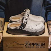 A pair of white cloud-style clogs with black speckles and silver buckle straps, displayed on top of a brown shoebox branded MADEN ZEPPELIN CLOUD SLIPPERS. The background shows a person wearing a blue jacket, making this a lifestyle/product presentation shot.