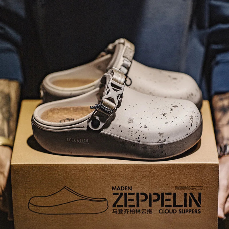 A pair of white cloud-style clogs with black speckles and silver buckle straps, displayed on top of a brown shoebox branded MADEN ZEPPELIN CLOUD SLIPPERS. The background shows a person wearing a blue jacket, making this a lifestyle/product presentation shot.