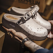 A pair of white clog-style shoes with black speckled detailing and a dark gray strap, held in hands in a warm, cardboard-filled environment. The brand name 'LOCK & TECH' is visible on the side of the shoe.