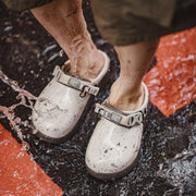 A close-up of a person's legs and feet wearing white speckled rubber clogs with metal buckle straps, standing in splashing water on a wet dark surface with orange-red painted sections visible.