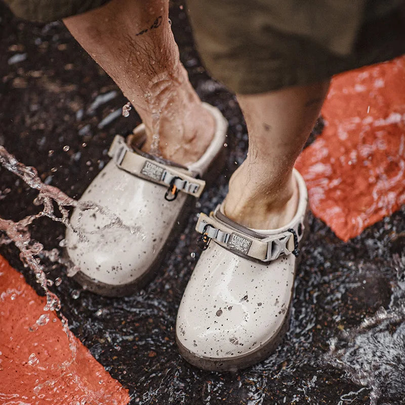 A close-up of a person's legs and feet wearing white speckled rubber clogs with metal buckle straps, standing in splashing water on a wet dark surface with orange-red painted sections visible.